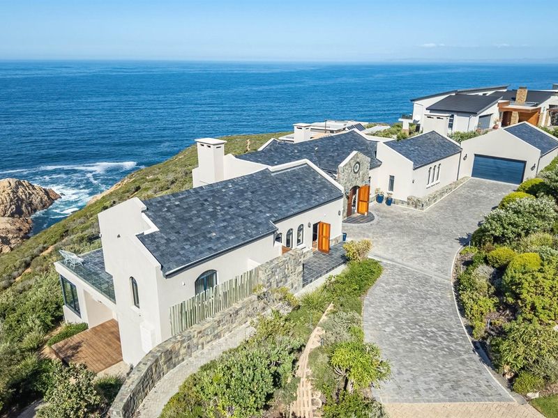 Aerial view of a luxury coastal residence featuring slate roofing, stone accents, a paved driveway, and expansive ocean views.