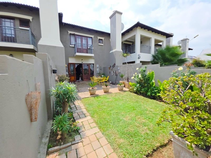 A private rear garden featuring a paved patio area, a well-maintained lawn, and direct access to the townhouse via timber-framed doors and upper-level balconies.