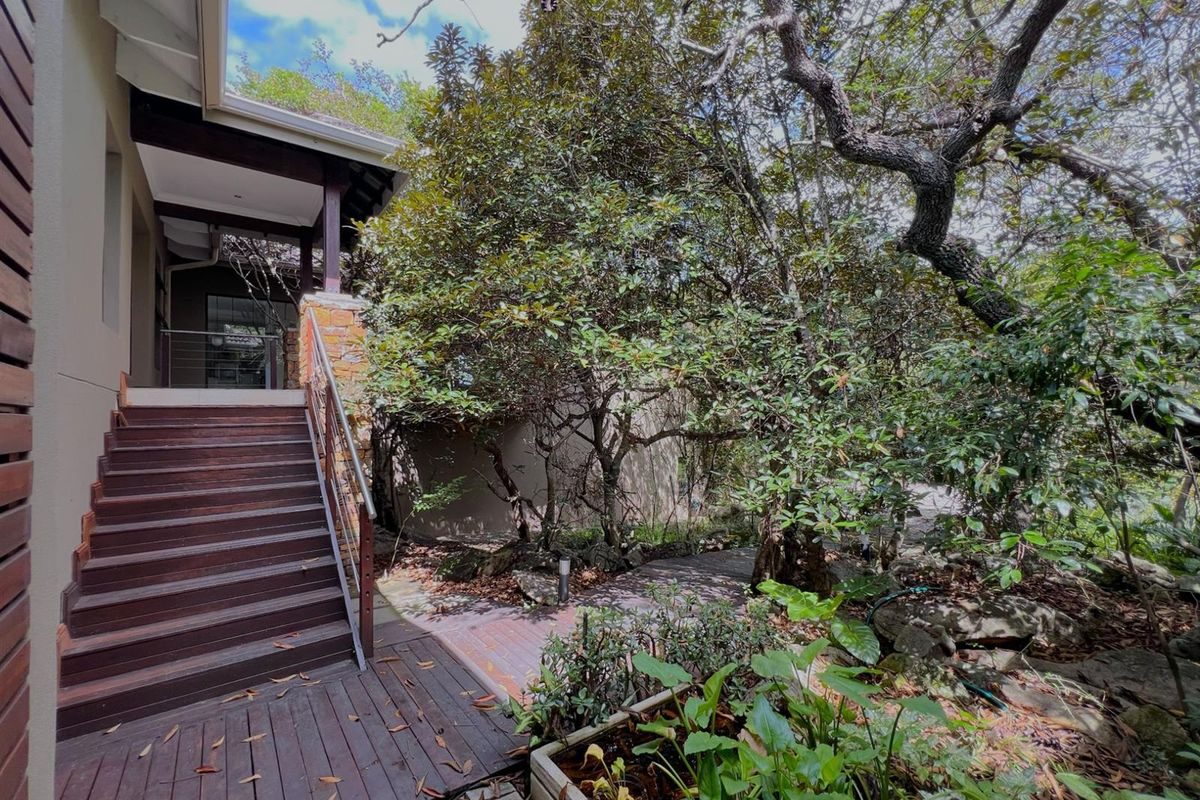 Rear exterior view featuring a timber staircase leading to a covered patio area, surrounded by mature trees and landscaped garden pathways.