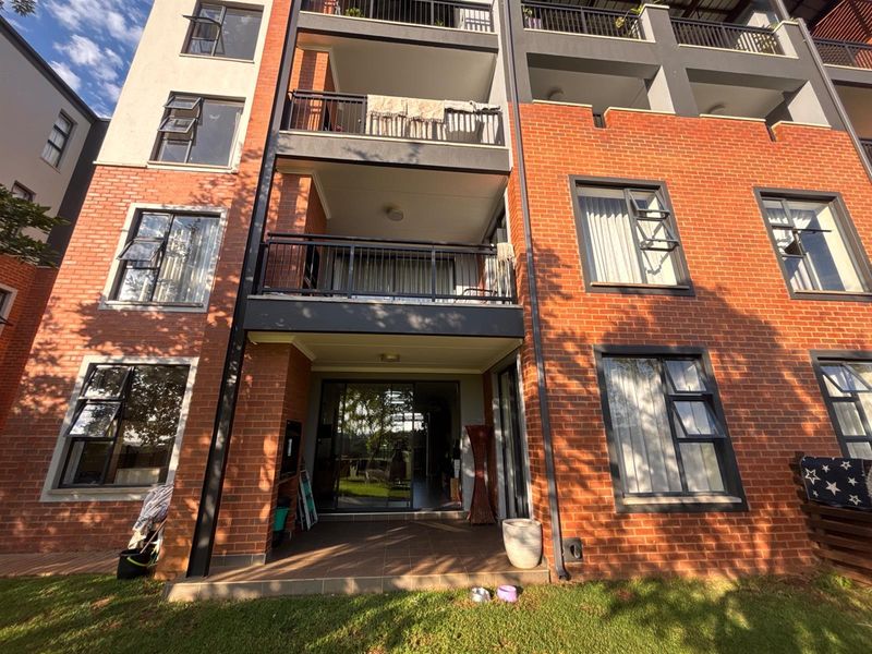 Rear view of a multi-story apartment building featuring red brick facade, private balconies with metal railings, and ground-floor patio access.