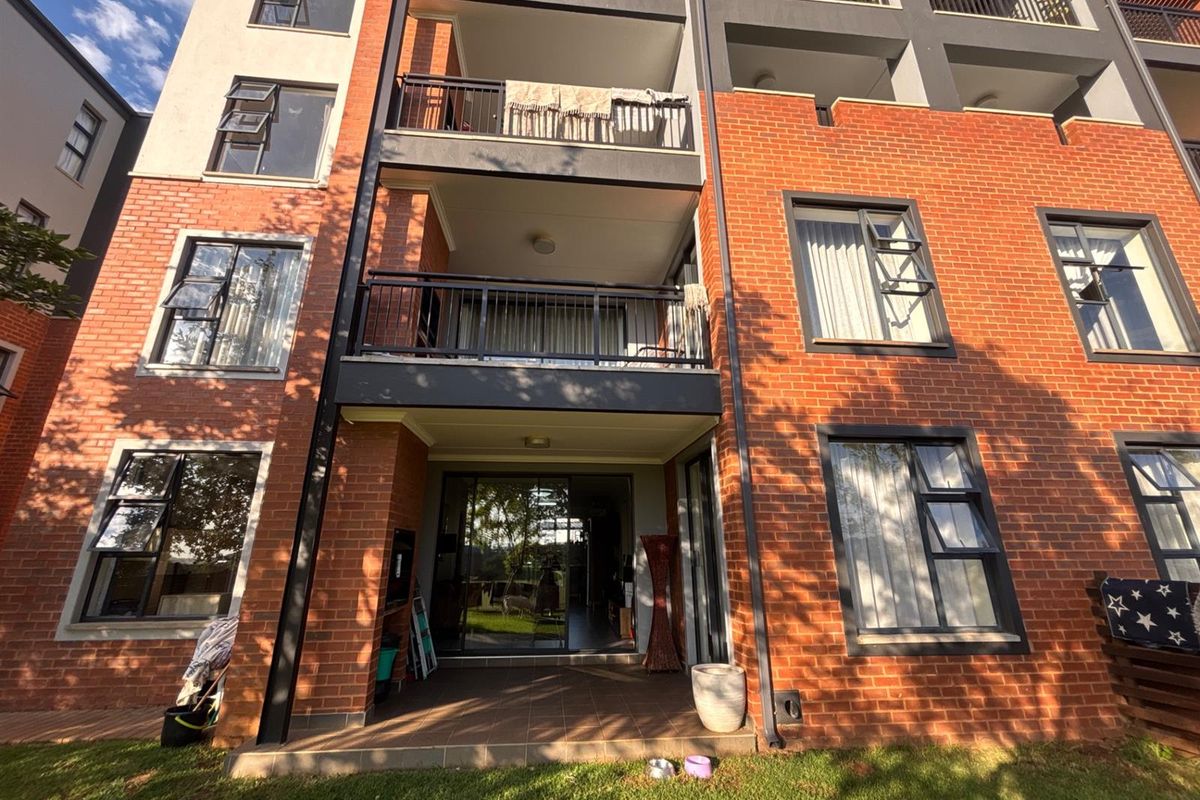 Rear view of a multi-story apartment building featuring red brick facade, private balconies with metal railings, and ground-floor patio access.
