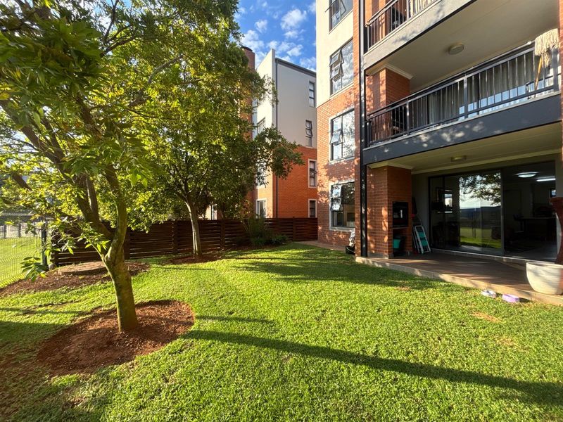 A well-maintained rear garden area featuring a lawn, mature trees, and a ground-floor patio with access to the apartment via sliding glass doors.
