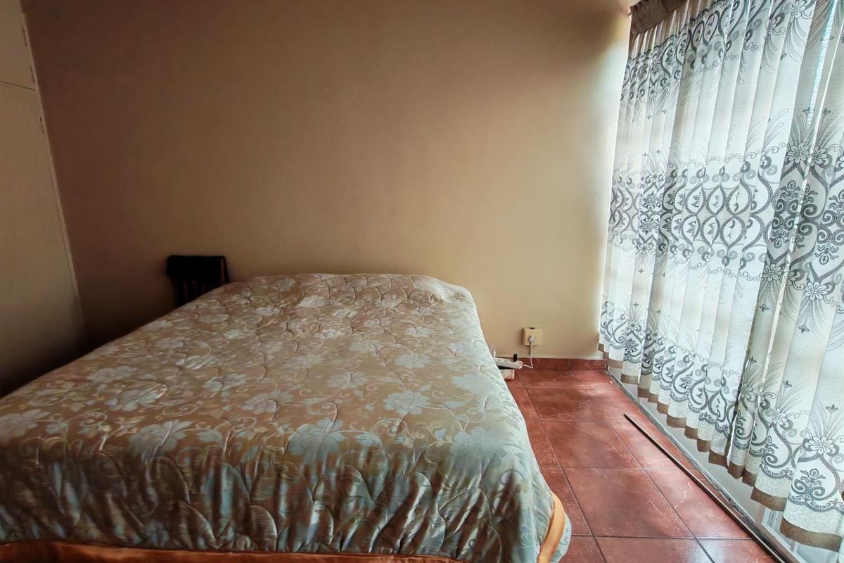 A bedroom featuring terracotta floor tiles and large windows with patterned curtains, providing natural light to the space.