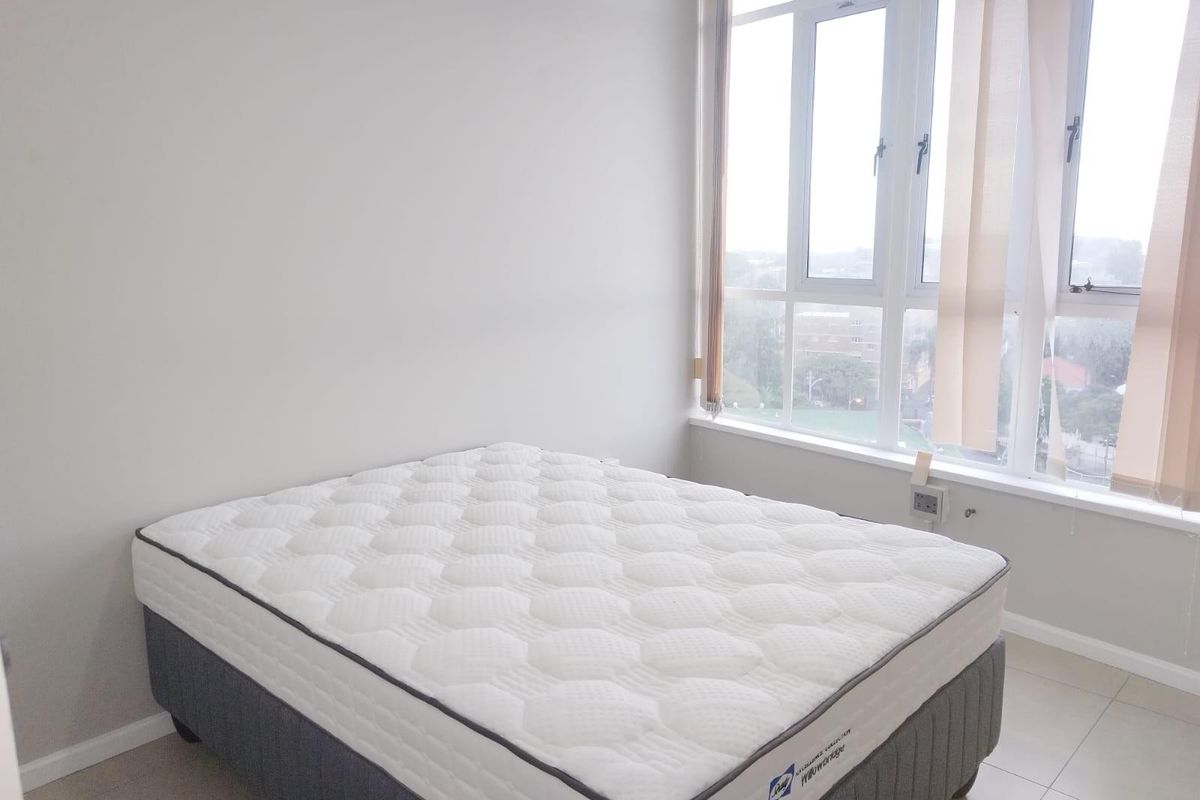 A bright, minimalist bedroom featuring large windows with natural light, neutral wall tones, and tiled flooring.