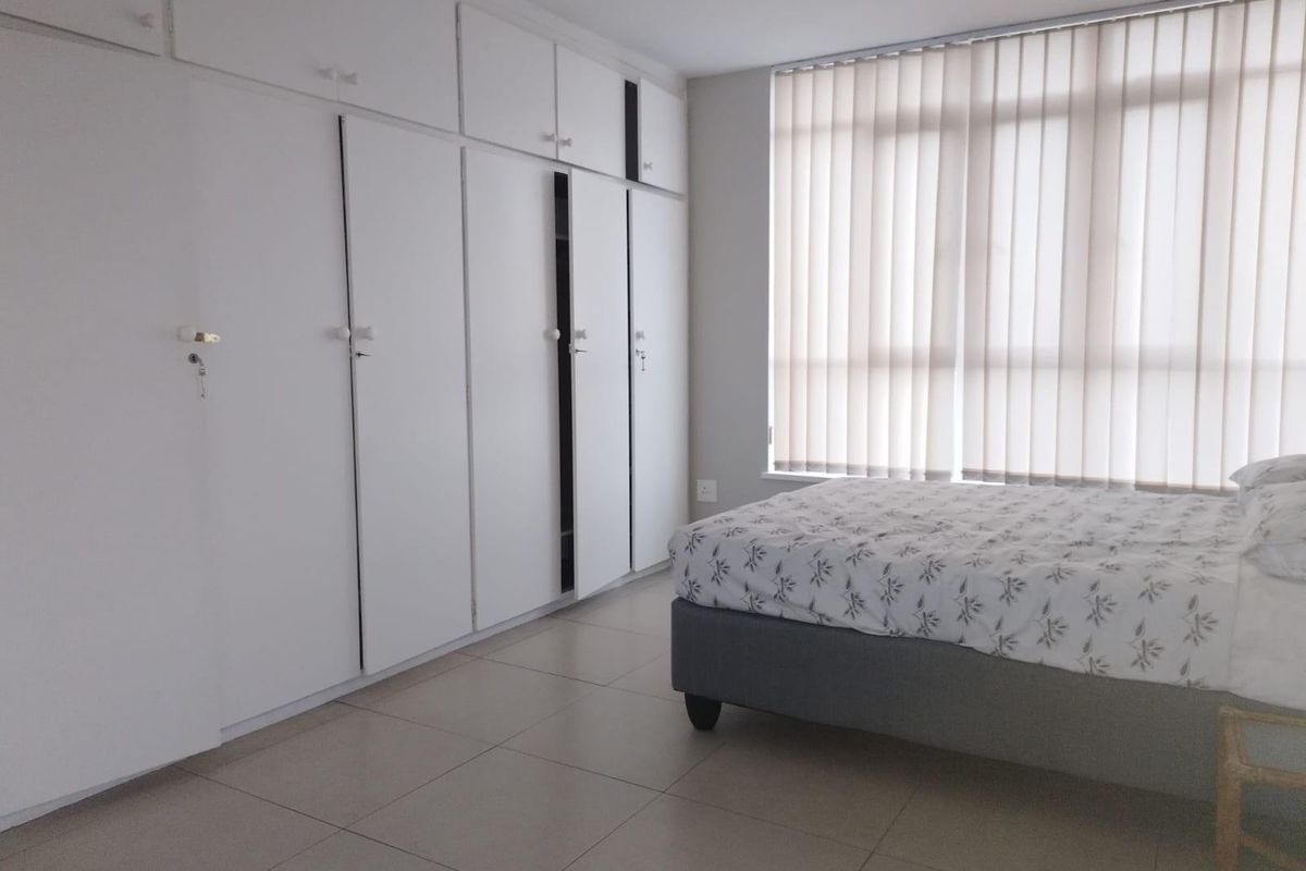 A bedroom featuring neutral tiled flooring, extensive white built-in cupboards, and vertical window blinds providing natural light.