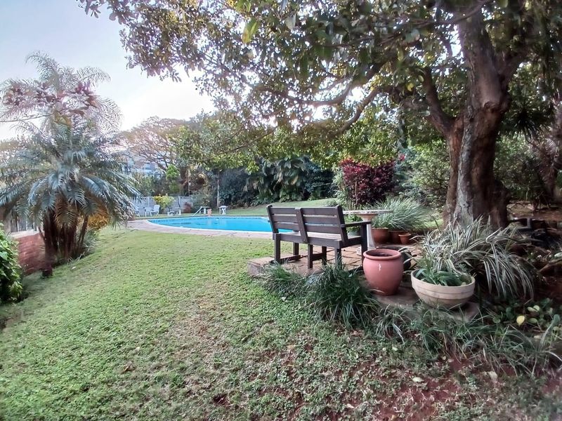 A lush garden setting featuring a swimming pool with paved surround and mature trees, providing a tranquil outdoor space.