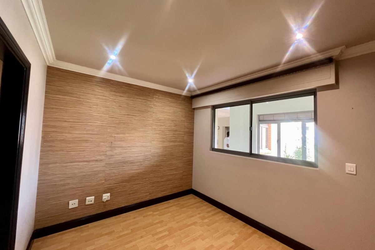 A bright, empty bedroom featuring wood-look laminate flooring, a textured accent wall, recessed ceiling lighting, and a sliding window with a roller blind.