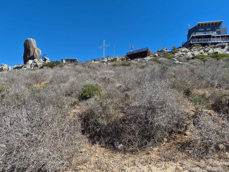 A view of the sloped, rocky terrain featuring native vegetation, an elevated wooden deck, and a large metal cross against a clear blue sky.