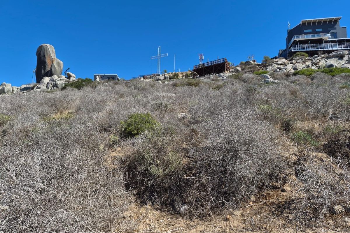 A view of the sloped, rocky terrain featuring native vegetation, an elevated wooden deck, and a large metal cross against a clear blue sky.