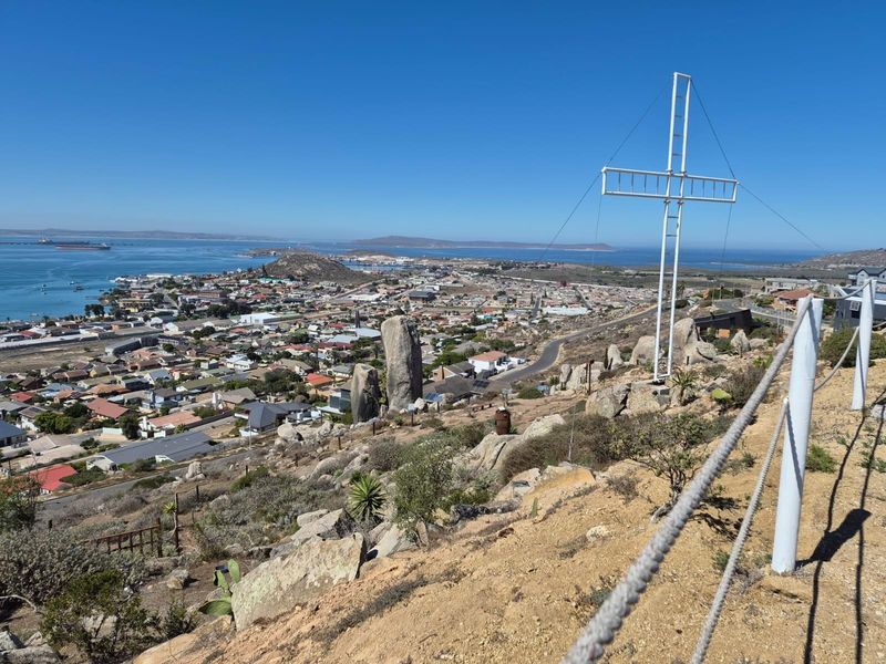 Elevated panoramic view overlooking the coastal town and bay, featuring a prominent metal cross structure and safety rope railing on the hillside.