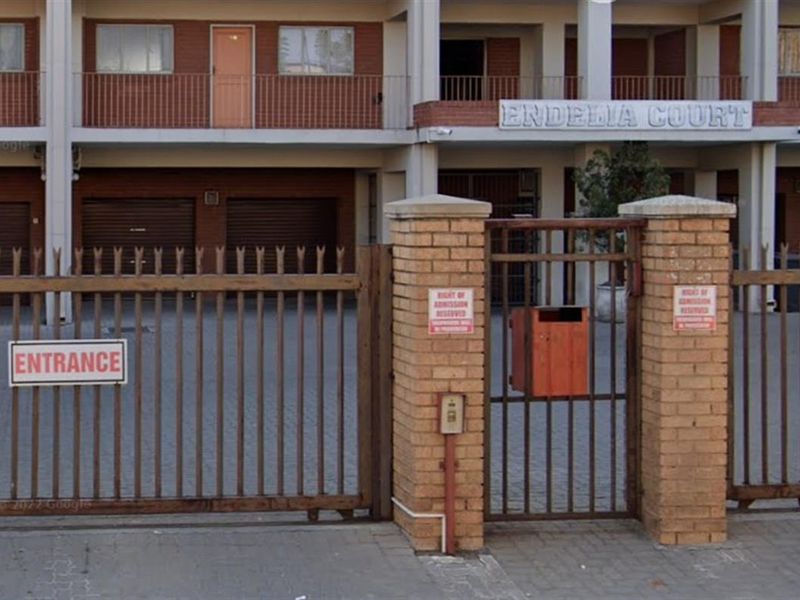 Front entrance view of Endelia Court apartment complex featuring secure gated access, brick boundary walls, and ground-level roller garage doors.