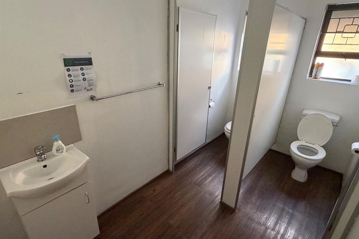A functional office bathroom featuring a vanity basin, toilet, and durable laminate flooring with natural light from a window with security bars.