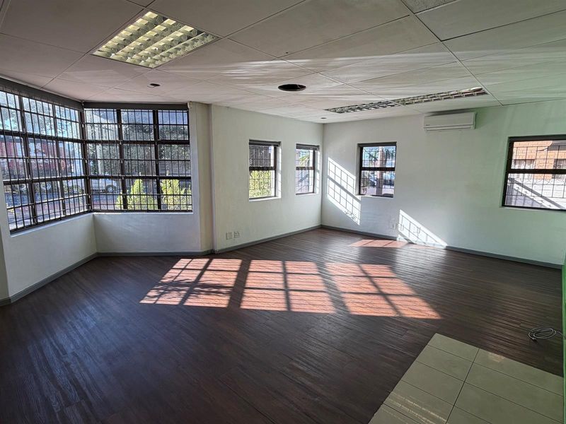 A bright, vacant office space featuring laminate flooring, security-barred windows, and a suspended ceiling with integrated lighting and air conditioning.