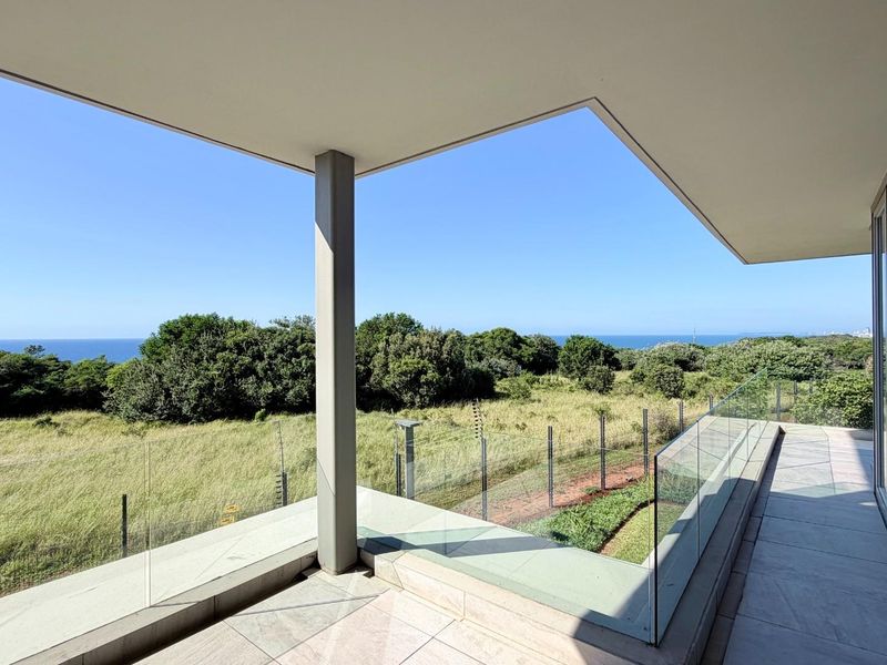 A spacious covered balcony featuring modern tiled flooring and a sleek glass balustrade, offering expansive views of the surrounding landscape and coastline.