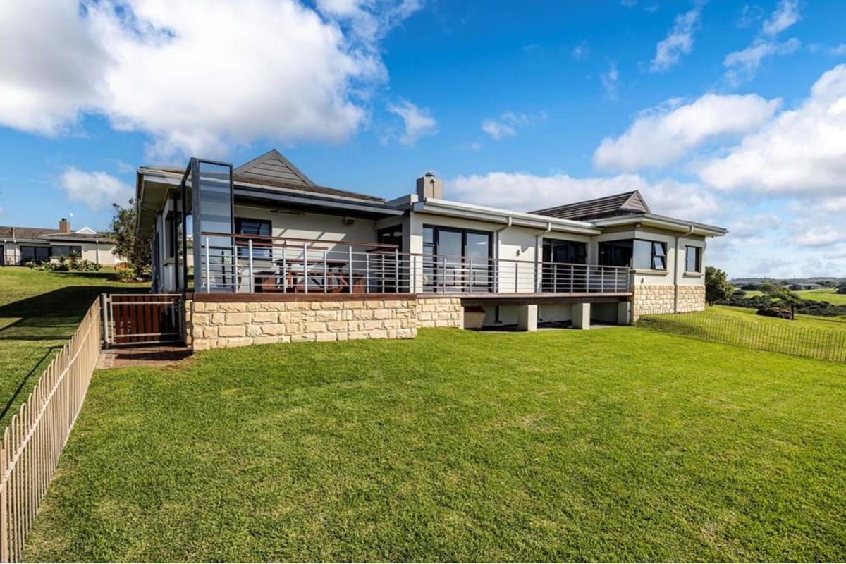 Rear view of the property featuring a raised timber deck with stainless steel balustrades, stone cladding, and large sliding glass doors overlooking the lawn.