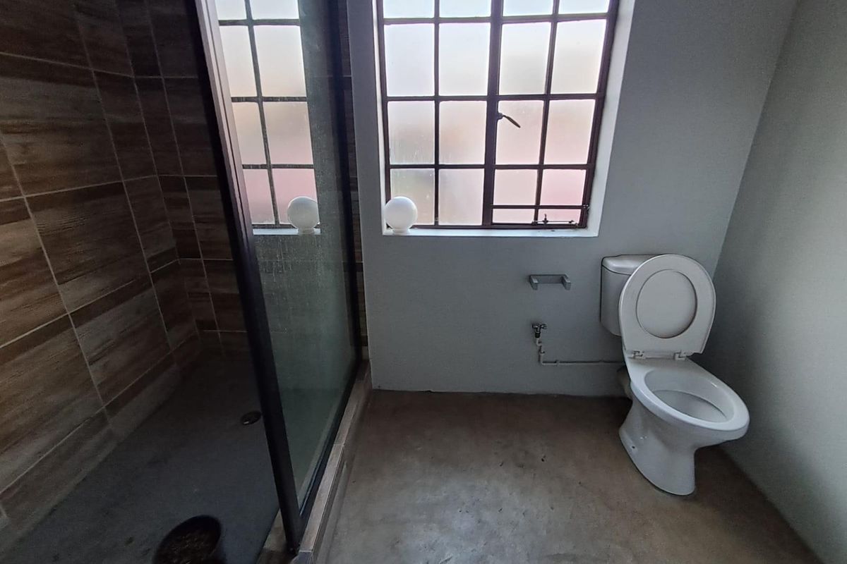 A functional bathroom featuring a glass-enclosed shower with wood-look wall tiling, a standard toilet, and a window with security bars.
