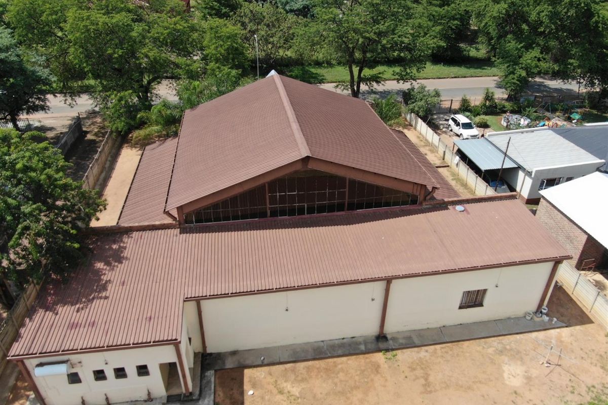 Aerial view of a commercial property featuring a large metal roof structure, surrounding grounds, and adjacent parking area.