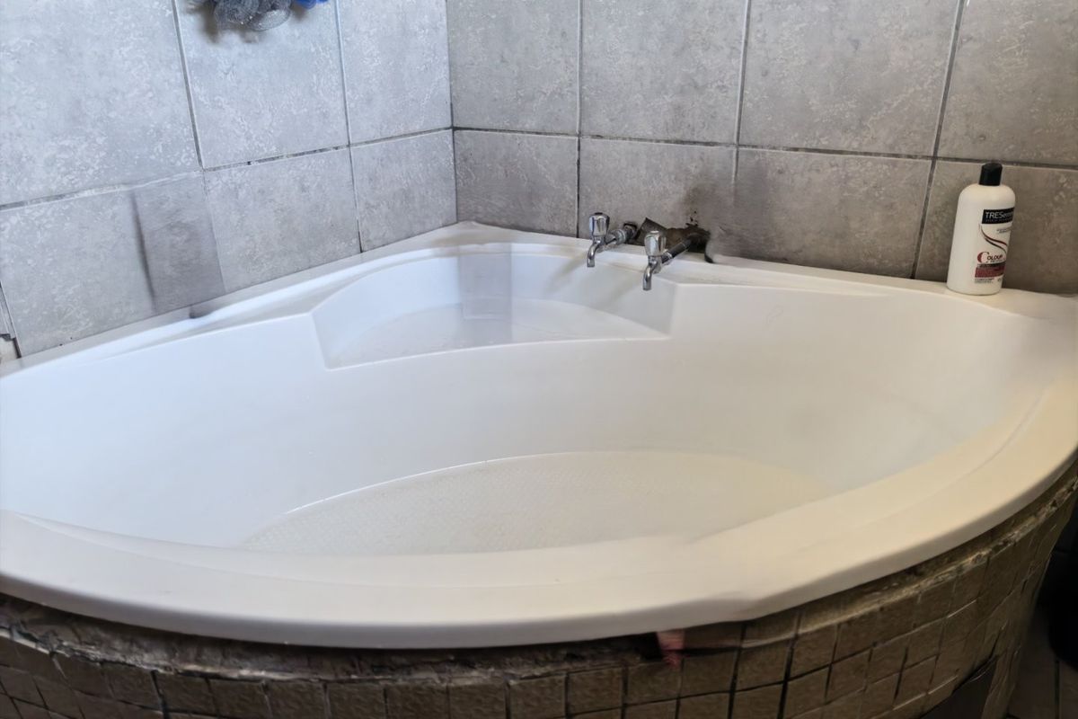 A corner bathtub featuring a mosaic tile apron and ceramic wall tiling, currently showing signs of wear around the faucet and base.