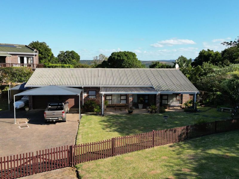 Aerial view of a single-story brick house featuring a corrugated metal roof, covered patio, paved driveway, carport, and a well-maintained garden with timber fencing.