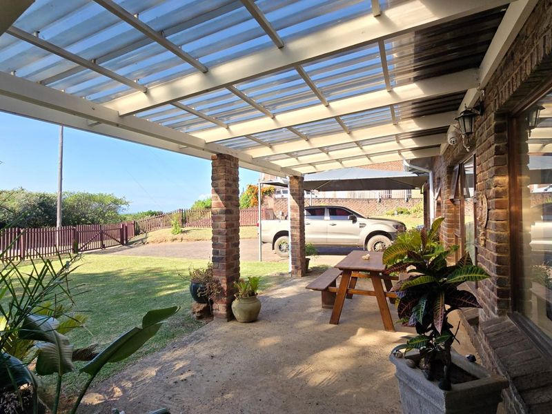 A covered outdoor patio area featuring brick pillars and a translucent corrugated roof, overlooking a private garden and driveway.