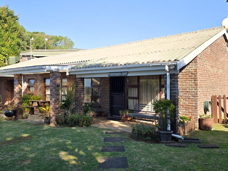 A single-story face brick house featuring a covered front patio, corrugated metal roof, and a well-maintained garden with a paved walkway.