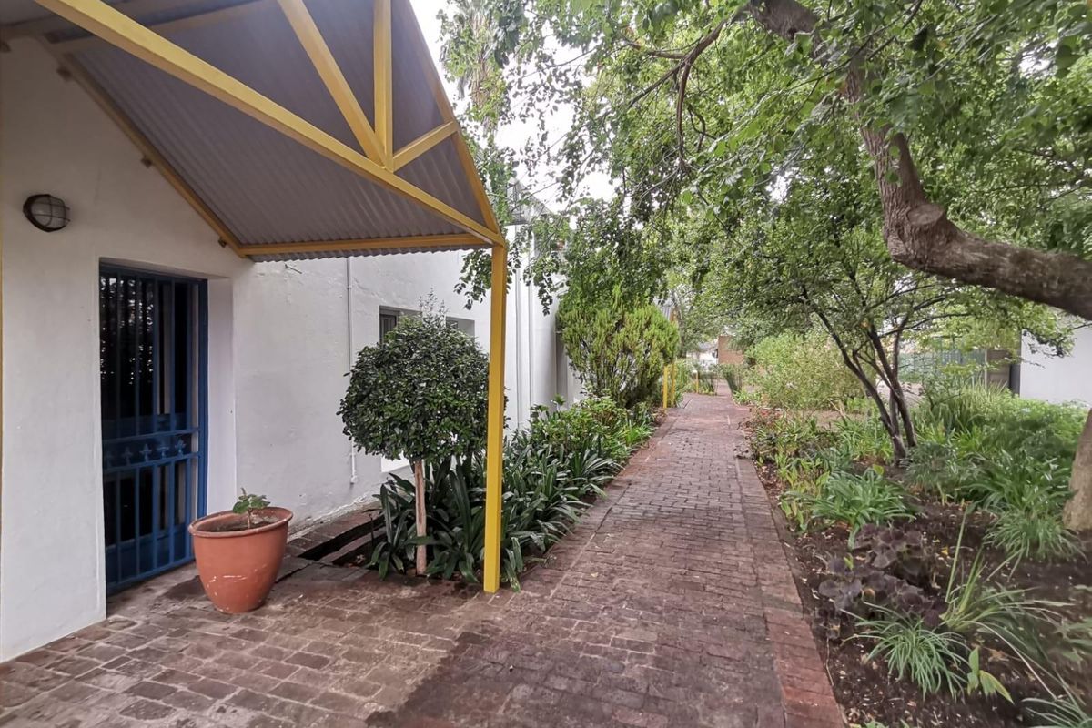 A paved walkway leads along the side of the office building, featuring a covered entrance with a yellow metal frame and established garden landscaping.
