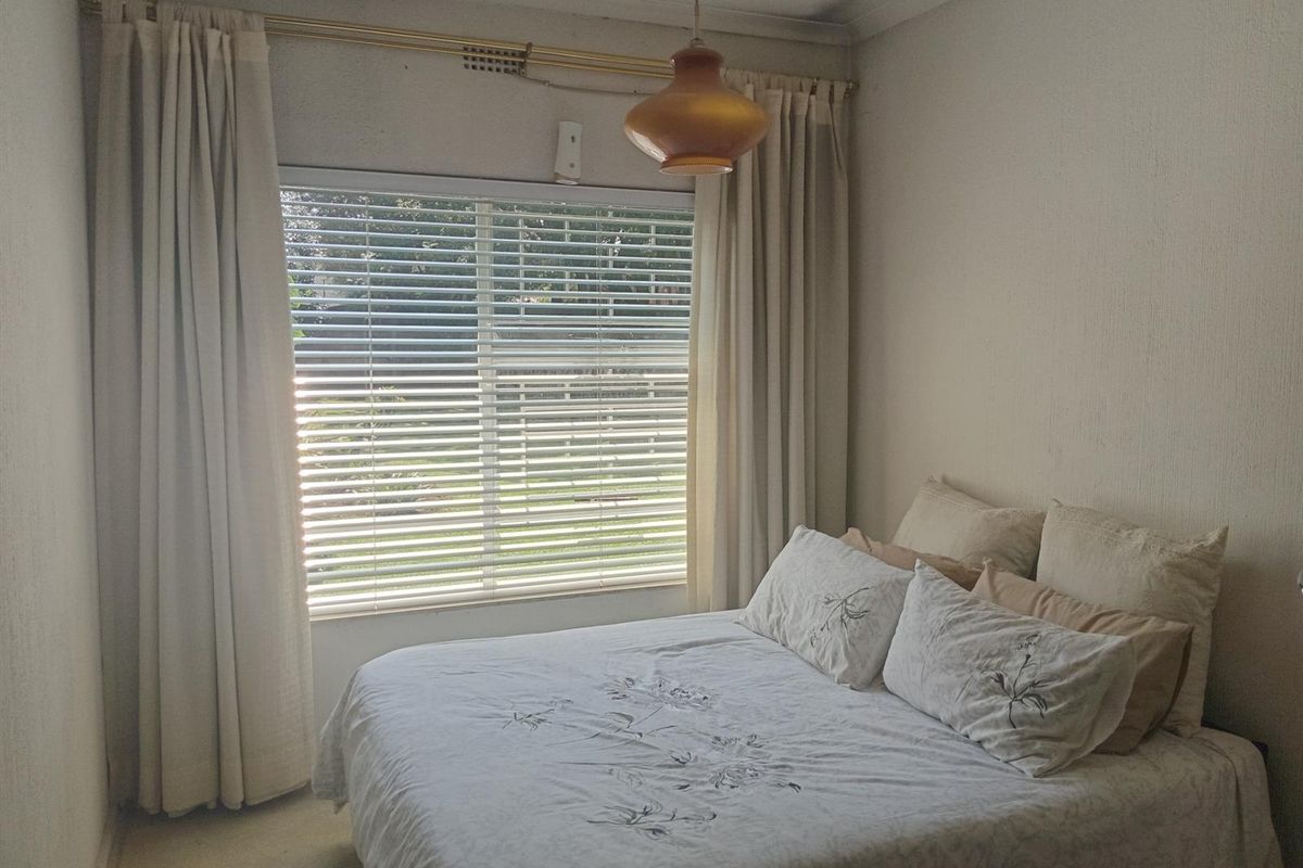 A functional bedroom featuring a large window with venetian blinds, neutral wall finishes, and a central pendant light fixture.