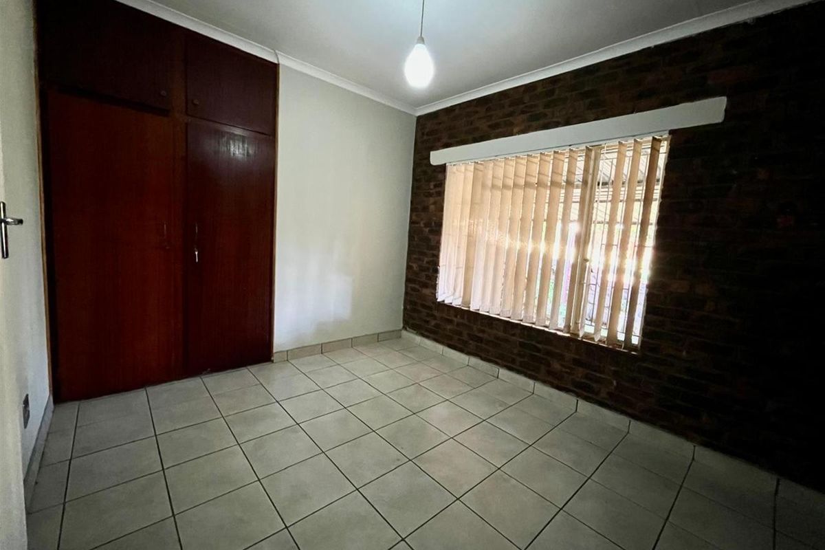 A vacant bedroom featuring tiled flooring, built-in cupboards, and an exposed brick feature wall with vertical blinds.