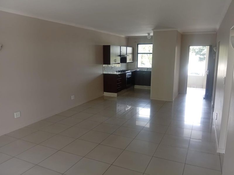 An open-plan living and kitchen area featuring neutral tiled flooring, dark built-in cabinetry, and integrated kitchen appliances in a clean, vacant space.
