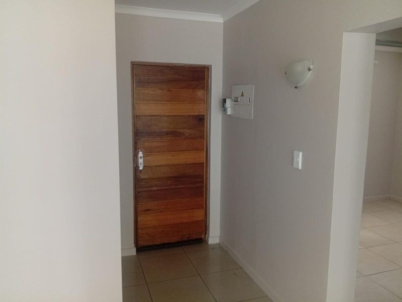 A clean and simple apartment entrance featuring a solid wooden door, neutral tiled flooring, and a wall-mounted electrical distribution board.