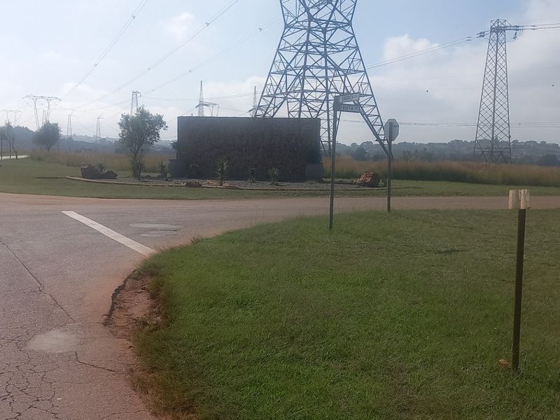 Street view of a vacant residential land plot featuring a stone entrance monument, paved road access, and overhead high voltage power lines.
