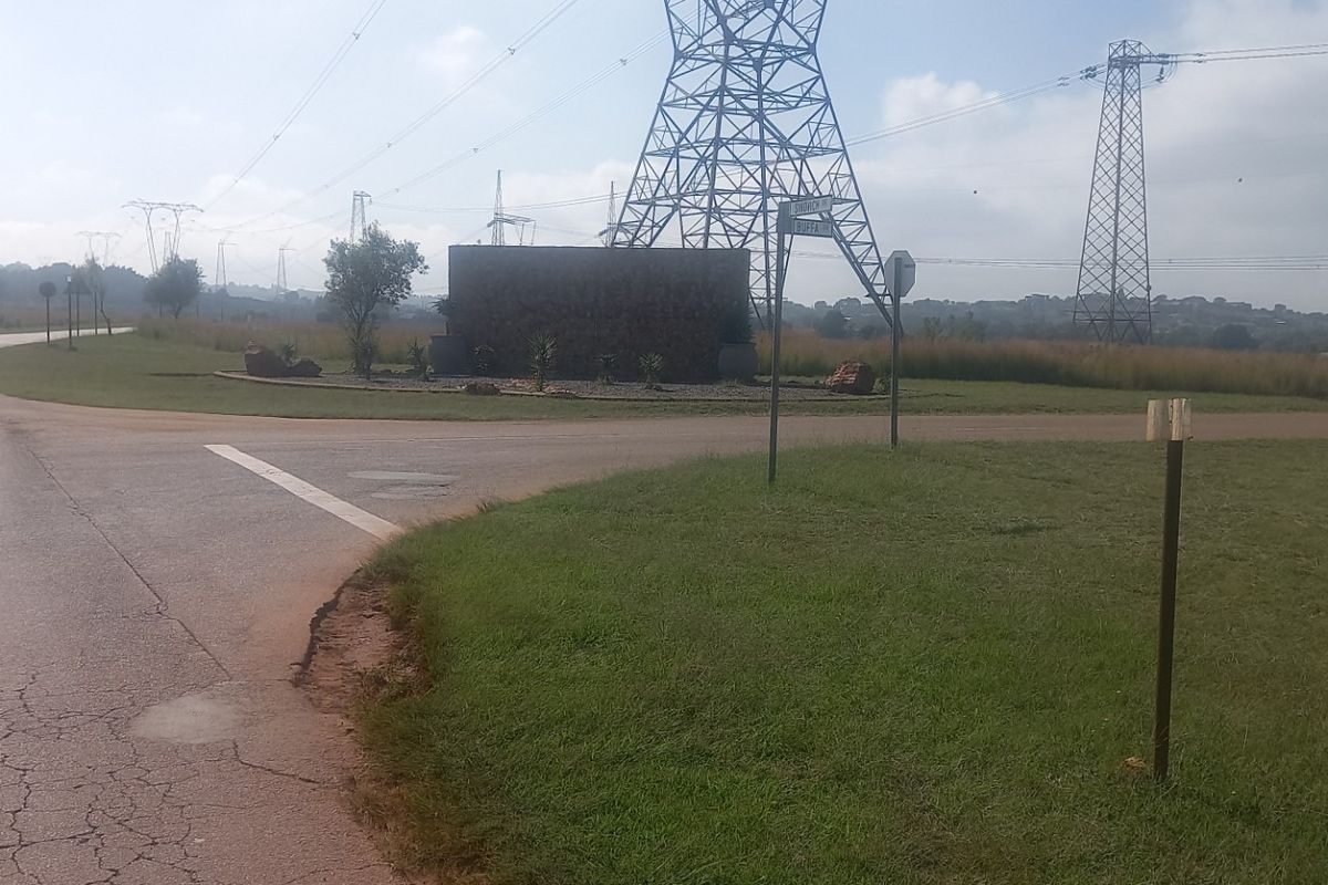 Street view of a vacant residential land plot featuring a stone entrance monument, paved road access, and overhead high voltage power lines.