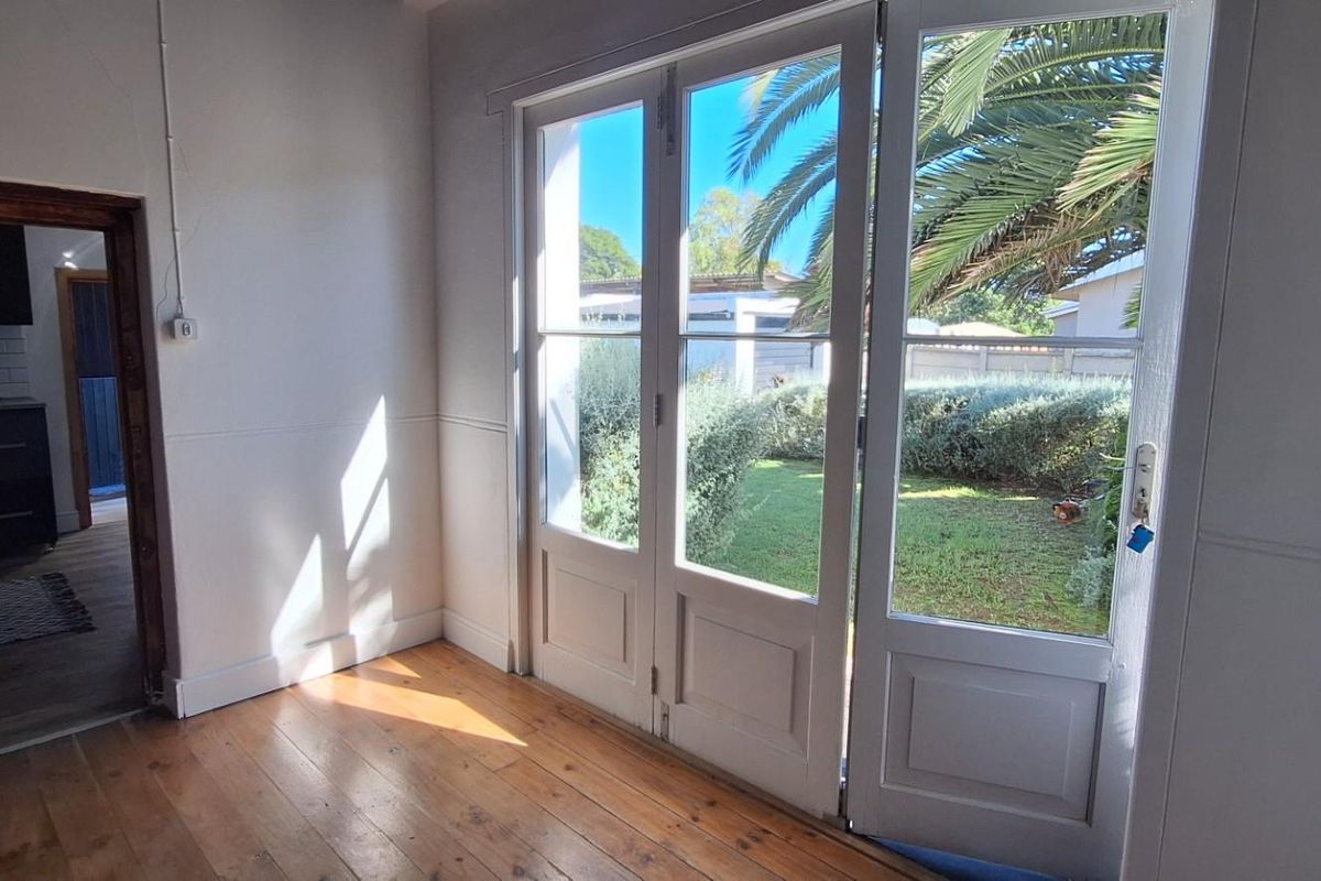 Bright living area featuring polished timber flooring and double glass doors opening onto a private garden, with a view into the adjacent kitchen.
