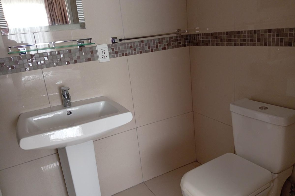 A clean bathroom featuring a white pedestal basin, a modern toilet, and neutral wall tiles with a decorative mosaic border.