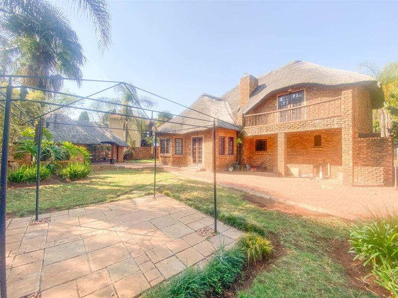 Rear view of a double-story brick house with a traditional thatch roof, featuring a paved patio area and a metal pergola frame in the garden.