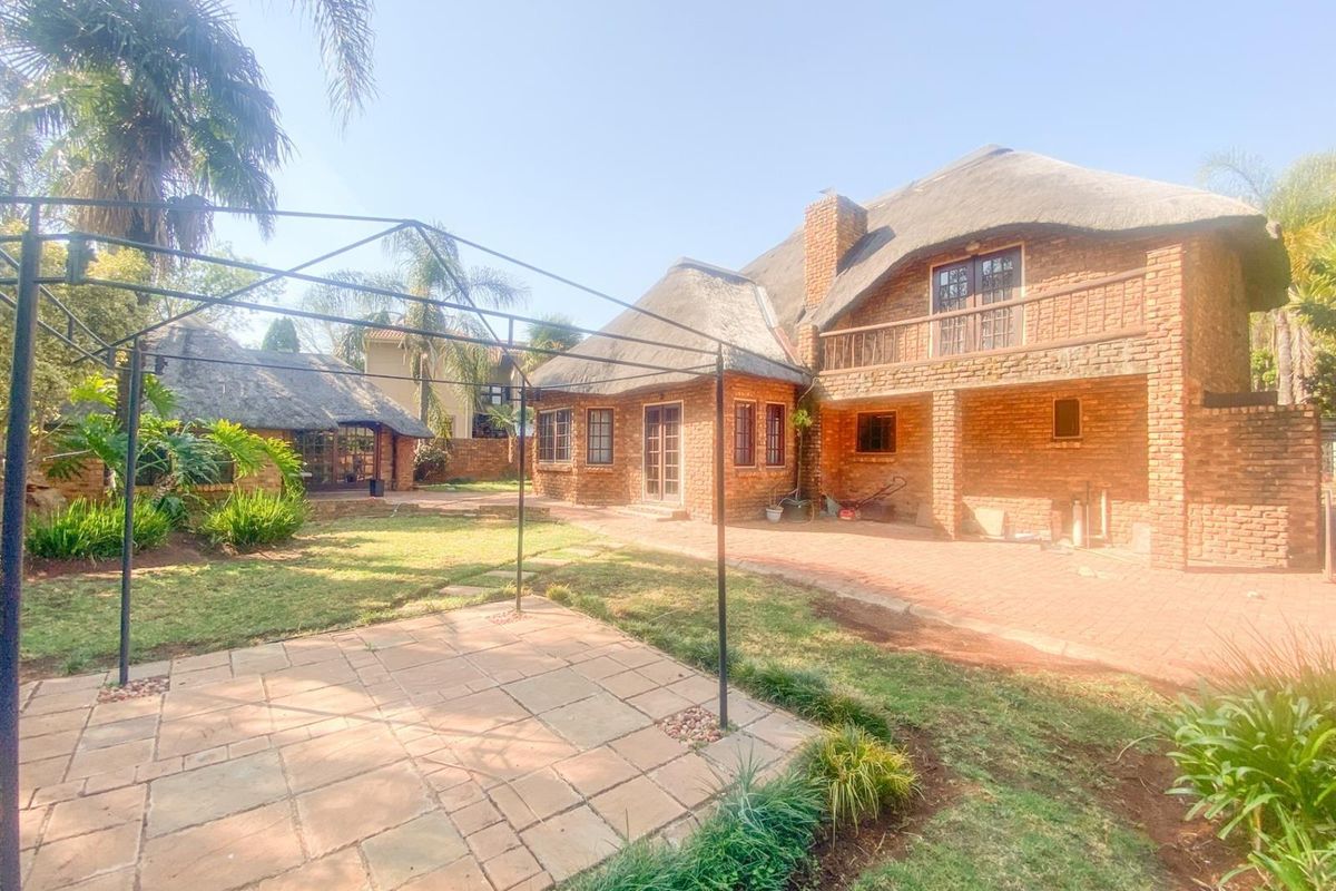 Rear view of a double-story brick house with a traditional thatch roof, featuring a paved patio area and a metal pergola frame in the garden.