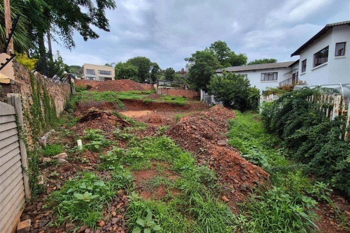 A vacant residential plot featuring uneven terrain, significant earthworks, and overgrown vegetation, bordered by existing residential properties and boundary walls.