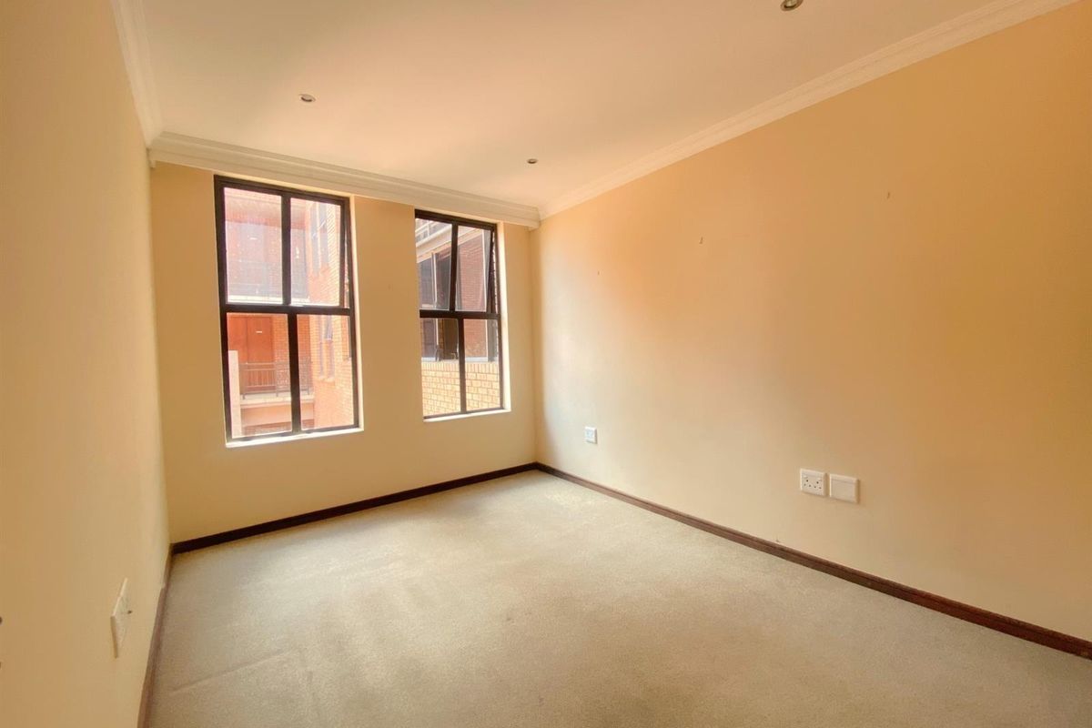 A bright, empty bedroom featuring neutral wall tones, carpeted flooring, and large windows providing natural light and views of the surrounding apartment complex.