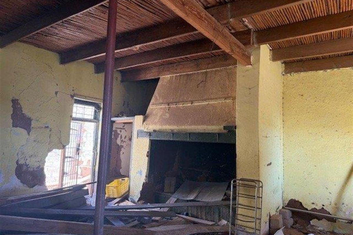 A dilapidated living room featuring a large built-in fireplace, exposed timber beam ceiling, and significant wall damage requiring extensive renovation.