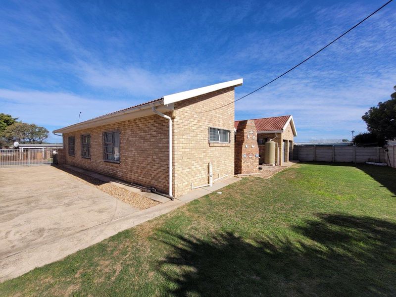 Rear view of the property featuring a brick facade, built-in braai, water tank, and a spacious lawn area with concrete driveway.