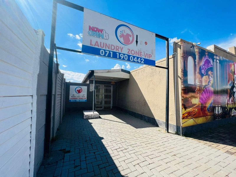 Exterior view of a commercial laundry facility entrance featuring paved access, security gate, and prominent business signage.
