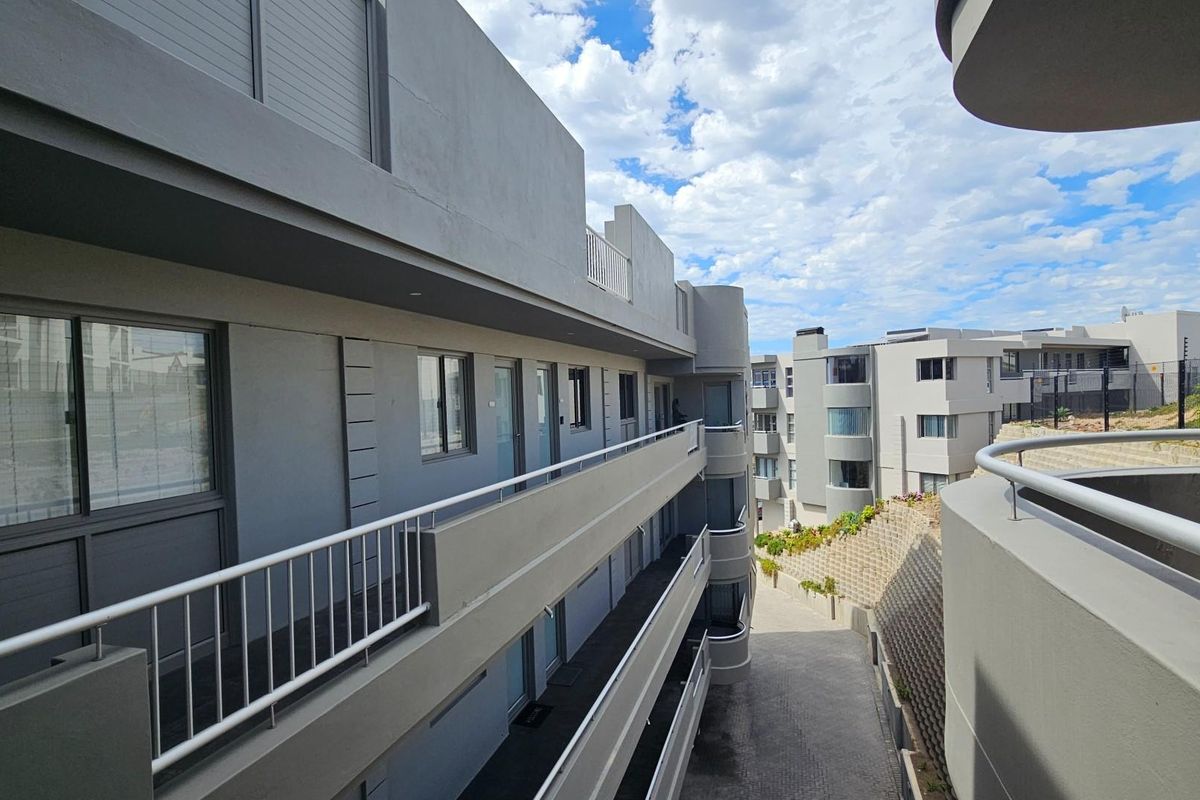 Modern apartment complex exterior featuring multi-level walkways, contemporary grey facade, and metal railings overlooking a paved driveway and retaining wall.