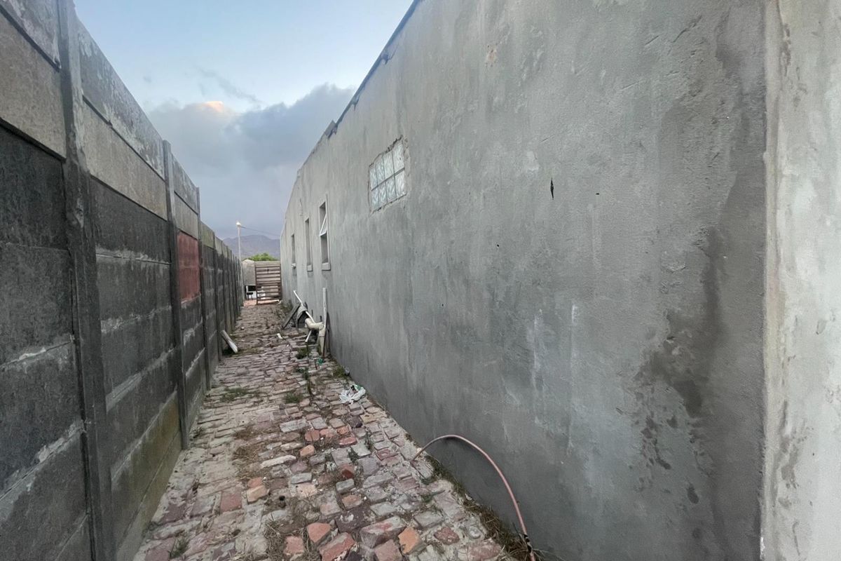 A narrow side passage featuring a brick paved walkway, unfinished exterior wall, and precast concrete boundary fencing.