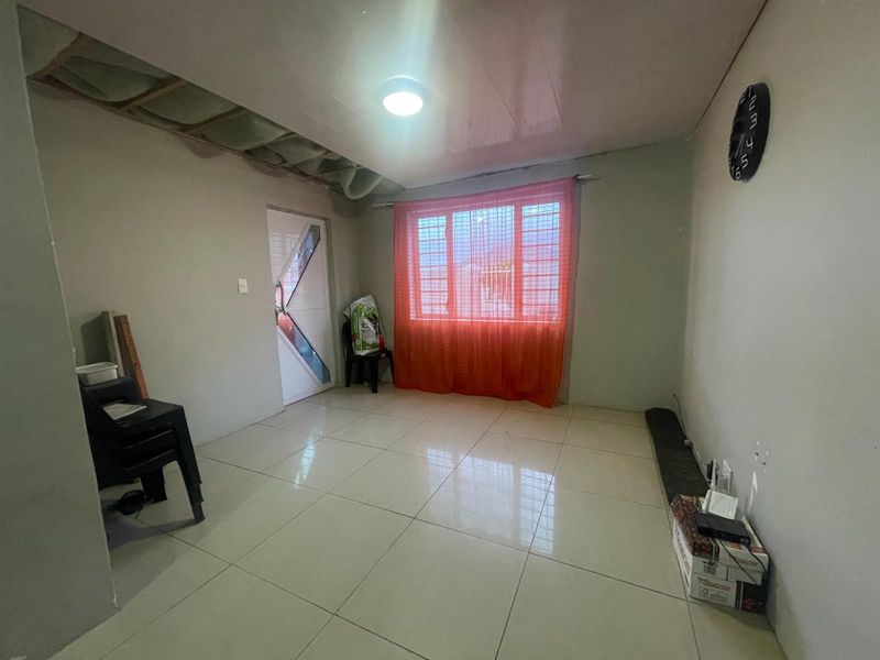 A sparsely furnished living area featuring tiled flooring, security window bars, and an unfinished ceiling section requiring completion.