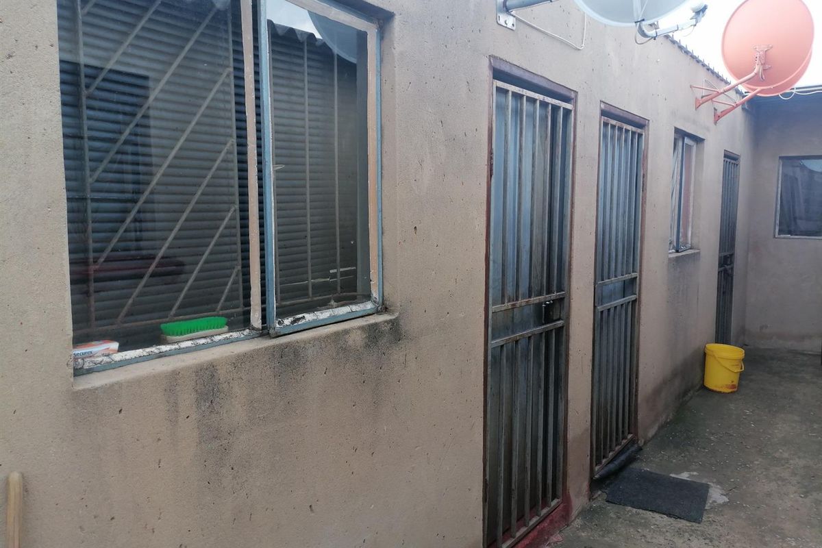 Rear exterior view of the property featuring concrete paving, security gates on doorways, and metal-framed windows with visible signs of weathering.