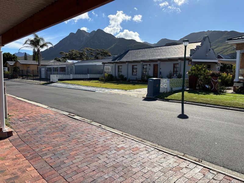 A clear street-level view of the property facade, featuring a gabled roof design, paved driveway, and mountain backdrop.