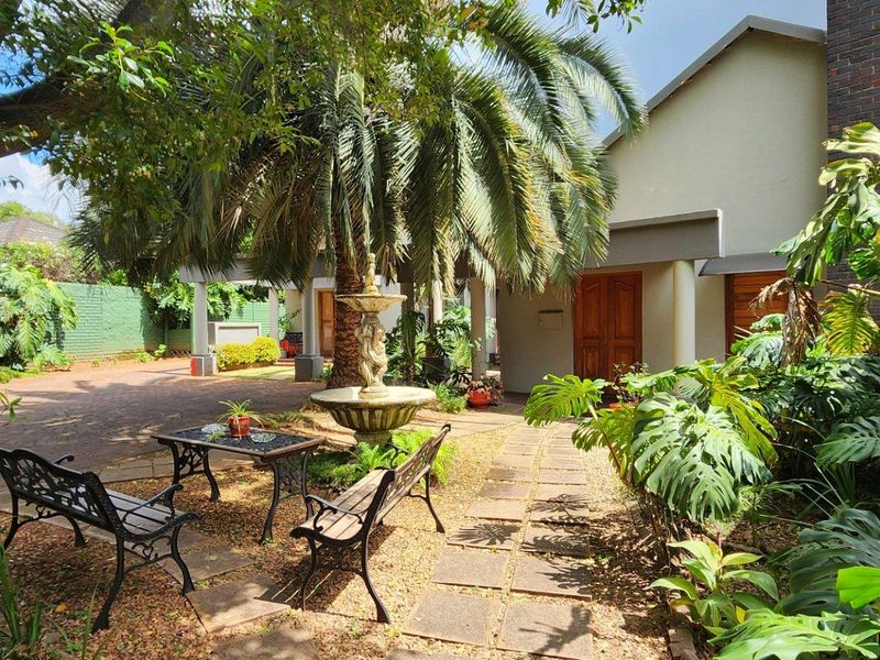 A landscaped front entrance area featuring a paved driveway, stone walkway, decorative fountain, and mature garden surroundings leading to the main building entrance.