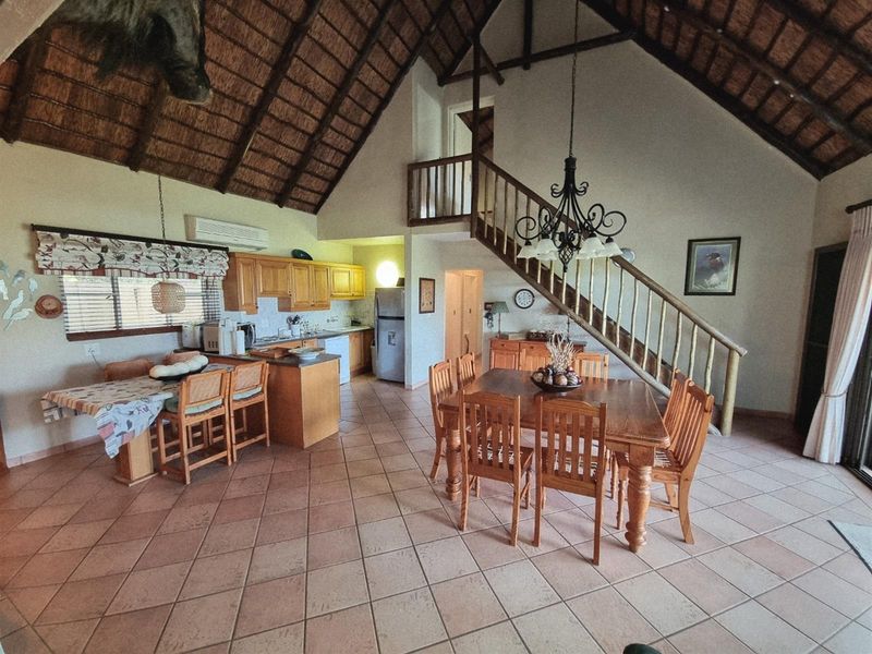 Open-plan living and dining area featuring a high thatched roof, exposed timber staircase, and integrated kitchen with wooden cabinetry.