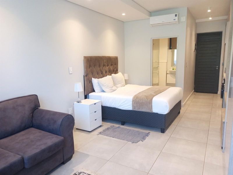 A modern bedroom featuring tiled flooring, recessed ceiling lighting, a wall-mounted air conditioning unit, and an en-suite bathroom entrance.