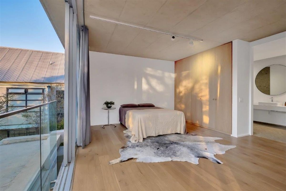 Modern bedroom featuring light timber flooring, exposed concrete ceiling, floor-to-ceiling built-in cabinetry, and a glass sliding door opening to a balcony.